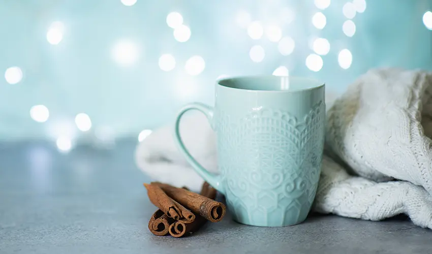 blue mug next to cinnamon sticks and a white blanket with a wintery background