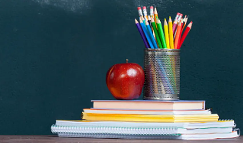An apple and a metal cup with colored pencils sits on top of a stack of books and spiraled notebooks